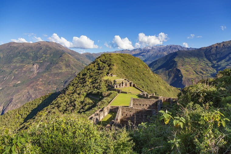 Ancient stone ruins surrounded by lush green hills and mountains under a bright blue sky with scattered clouds, likely depicting an archaeological site along the Choquequirao Trek in a remote, elevated region of Peru.
