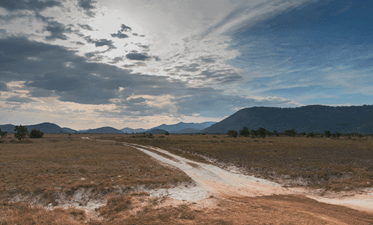 A dirt road runs through a dry, grassy plain under a partly cloudy sky, with mountains visible in the background.