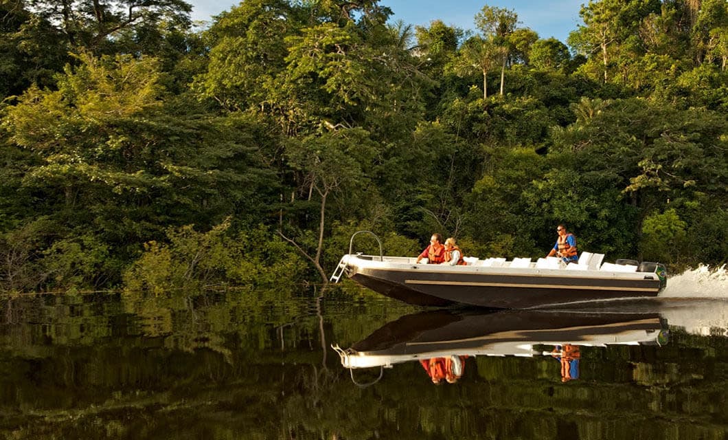 A motorboat with three people wearing life jackets speeds along a calm river during a Remote Amazon Tour, creating a reflection on the water. Dense green jungle trees cover the riverbank in the background.