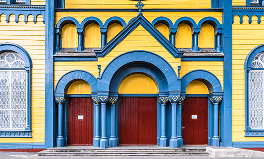 A bright yellow building with blue trim, arched red doors, decorative columns, and arched windows featuring white wrought iron designs—a vibrant stop on your Guianas Travel adventure tour.
