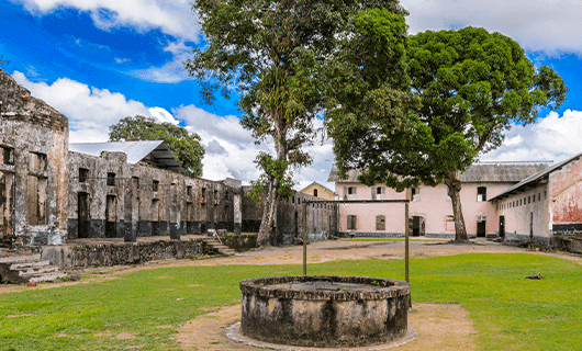 A circular stone well stands in the center of a grassy courtyard, surrounded by old, weathered stone buildings and large trees—a perfect stop on your Three Guianas adventure tour under a partly cloudy sky.