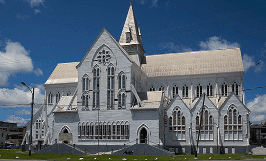 A large white wooden church with pointed arches and a tall central steeple stands under a blue sky, surrounded by grass and a few scattered birds—a striking landmark often admired on an Adventure Tour through the Three Guianas.