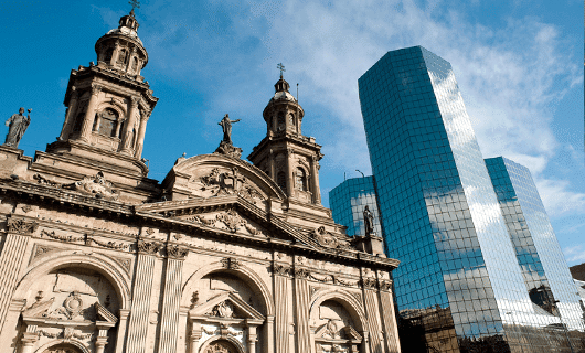 An old stone cathedral with ornate towers stands beside modern glass skyscrapers under a blue sky with scattered clouds, evoking the contrast seen during a Total Solar Eclipse in Chile.