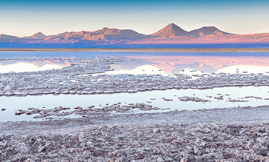 A tranquil salt flat in Chile with reflective pools of water in the foreground and distant mountains under a clear blue sky, bathed in soft early morning or evening light—an ideal setting for an unforgettable Eclipse Tour.
