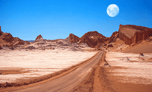 A straight dirt road cuts through a barren, rocky desert in Chile under a bright blue sky, with the full moon visible above distant jagged hills—an ideal setting for an unforgettable Eclipse Tour during a Total Solar Eclipse.
