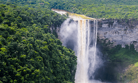 A large waterfall cascades over a cliff surrounded by dense, green rainforest, with mist rising where the water crashes below. Perfect for an Adventure Tour, the river flows at the top of the falls, cutting through the lush Three Guianas landscape.