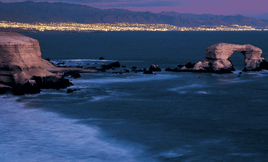 Rocky coastline at dusk in Chile with ocean waves, a large rock arch formation, and distant city lights glowing beneath a mountain range, all under a purple-blue sky perfect for an Eclipse Tour during the next Total Solar Eclipse.