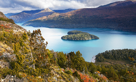 A scenic view of a turquoise lake in Chile, surrounded by forested mountains, with a small tree-covered island in the center and partly cloudy skies—perfect for an Eclipse Tour during a Total Solar Eclipse.