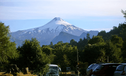 Snow-capped volcano rises above lush green forest in Chile, with parked cars and a bus visible in the foreground under a clear sky—a stunning scene for any Solar Eclipse Tour exploring the natural wonders of Chile.