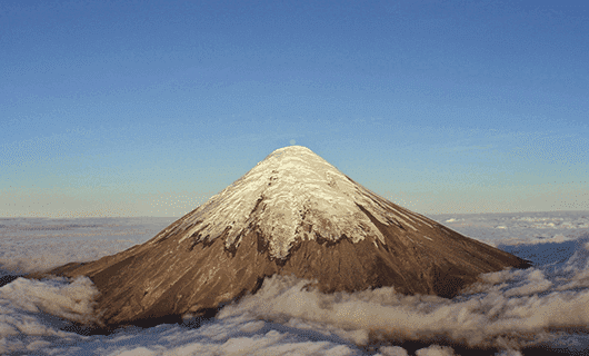A snow-capped volcanic mountain rises above a layer of clouds under a clear blue sky. Perfectly framed, its symmetrical summit makes an ideal backdrop for a Chile Eclipse Tour or witnessing the Chile Total Solar Eclipse.