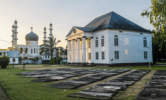 A white colonial-style building with columns stands near old stone graves, with a mosque featuring a gold dome and tall minarets in the background—an iconic sunset view perfect for any Guianas tour adventure.