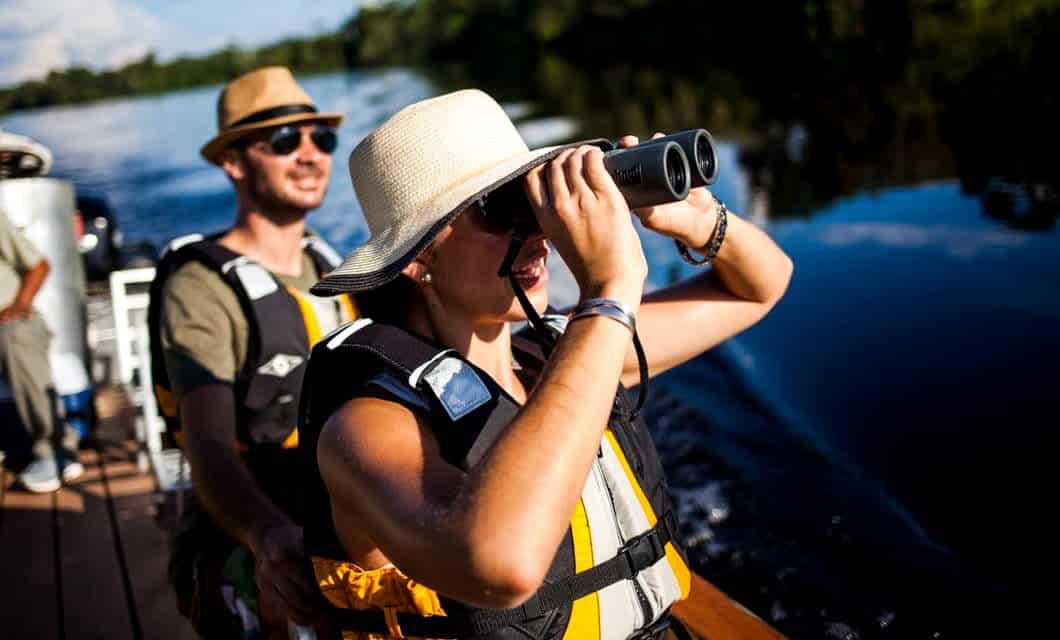 Two people in life jackets and hats stand on a boat during an Amazon Delfin II Cruise. The woman in front scans the scenery with binoculars while the man behind her smiles. The boat drifts along a calm river with trees in the background.