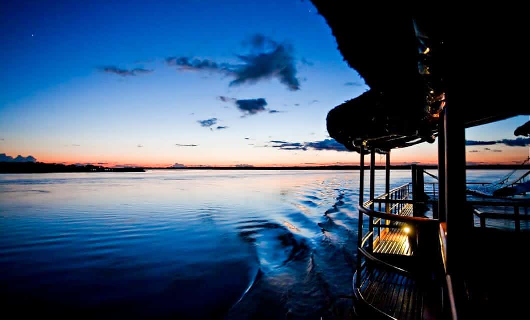 A boat cruises on a calm river at sunset, leaving gentle ripples in the water. The sky glows blue and orange as shimmering reflections drift by, evoking the serene beauty of an Amazon Delfin II Cruise.