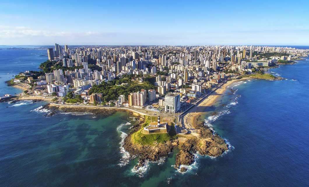 Aerial view of a coastal city with dense high-rise buildings, a curving sandy beach, green areas, and a prominent lighthouse on a rocky point—perfect for those seeking an urban tour before exploring the natural wonders of Brazil's Cerrado.