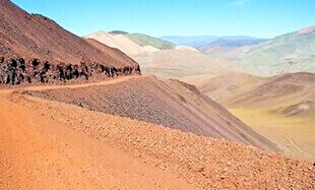 A narrow dirt road winds along the side of the barren, reddish-brown Velasco mountain in the Andes, overlooking a vast, arid landscape with rolling hills under a clear blue sky.