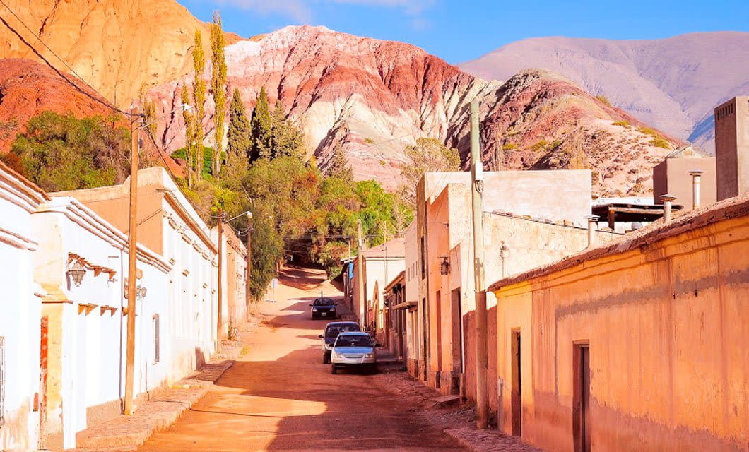 A narrow street lined with adobe-style buildings leads toward colorful, layered mountains under a blue sky. Cars parked along the road and trees add greenery—an inviting stop on any Coast to Coast Tour through this arid landscape.