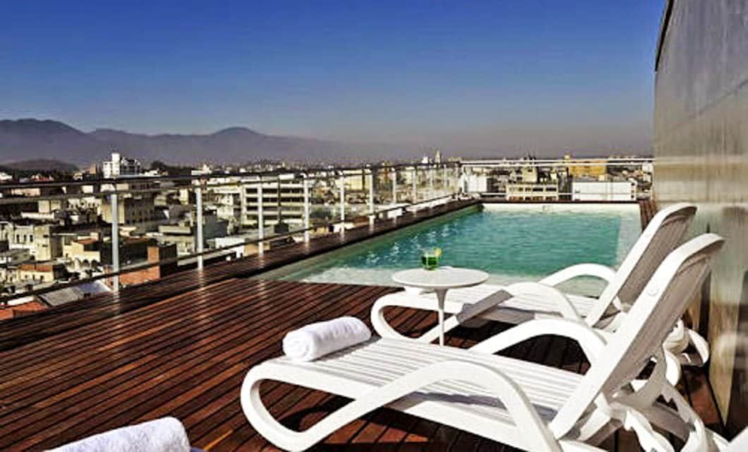 Rooftop pool area with white lounge chairs and a small table, overlooking a Salta cityscape with mountains in the background under a clear blue sky. Rolled towels and a drink are placed on the table and chairs.