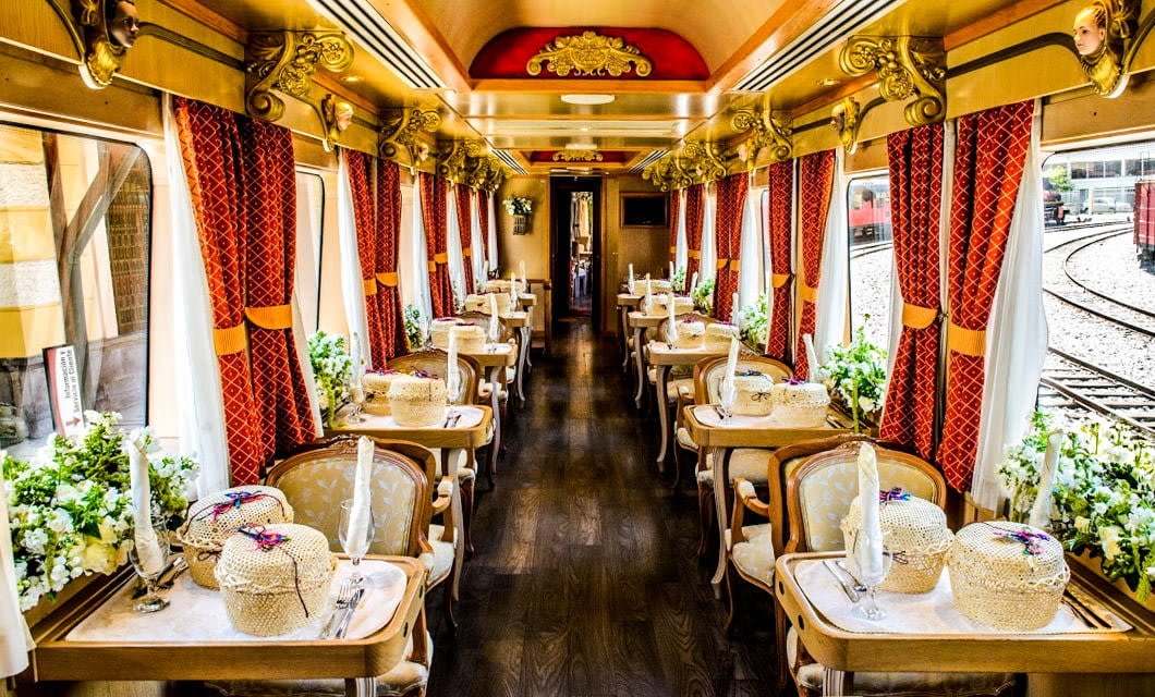 Elegant vintage train dining car with polished wood floors, red patterned curtains, and ornate ceiling recalls the refined ambiance of Ecuador Trains—tables set with wicker baskets and fresh flowers create an inviting, luxury travel experience.