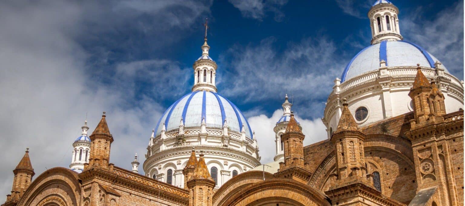 Close-up view of a historic cathedral in Ecuador with brown brickwork and three large blue-and-white domes under a bright, partly cloudy sky—a must-see highlight on any Ecuador Vacation.