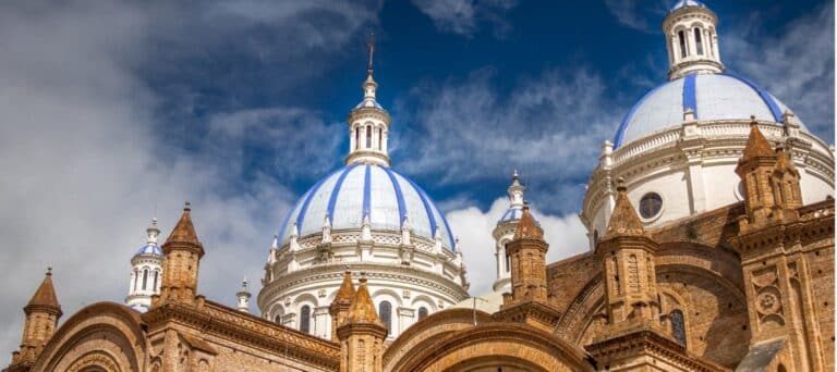 Close-up view of a historic cathedral in Ecuador with brown brickwork and three large blue-and-white domes under a bright, partly cloudy sky—a must-see highlight on any Ecuador Vacation.