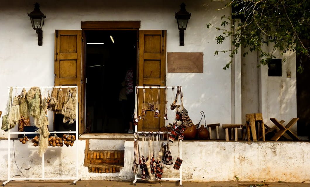 A rustic shop with wooden doors displays handmade crafts perfect for a Paraguay Tour—woven bags, necklaces, and artisanal goods stand outside. Wooden stools and gourds line the porch against a white wall, reflecting local charm.