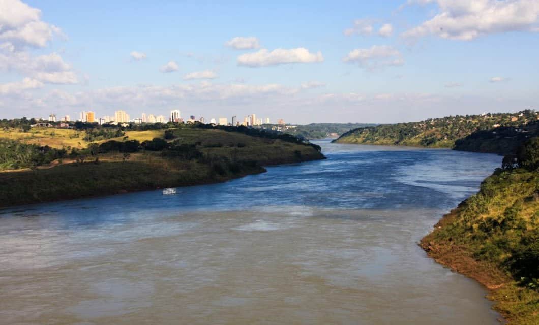 Wide river flowing between green, hilly banks under a partly cloudy sky, with a distant city skyline visible on the horizon and a small boat—perfect for a tranquil tour near Paraguay's border, reminiscent of scenes from Iguazu Falls.