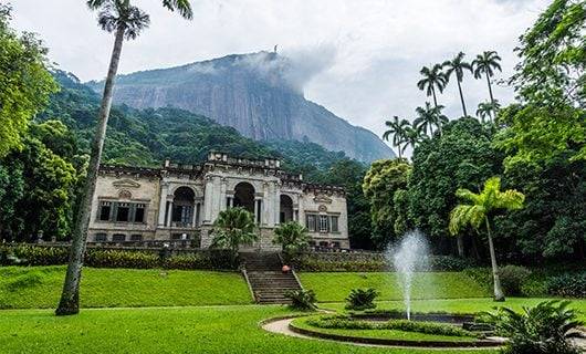 A historic mansion with arched windows and columns sits amidst lush greenery, framed by palm trees and a small fountain—an ideal spot to witness a Brazil solar eclipse with the misty mountain in the background.