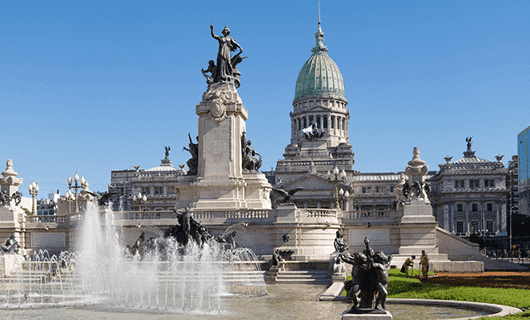A grand neoclassical building with a large green dome stands behind a monument and fountain, surrounded by statues and clear blue sky—an iconic sight on any Total Solar Eclipse Tour through Argentina.