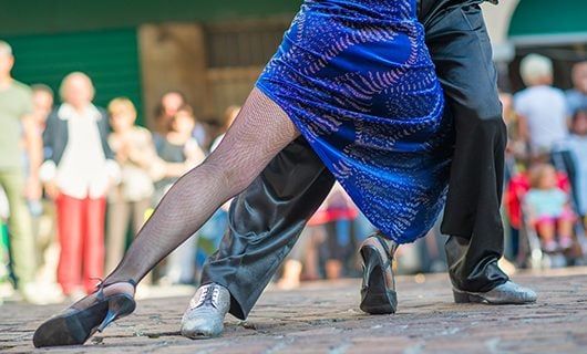 Two people dance closely on a cobblestone street in Argentina; the focus is on their legs. One wears a blue dress and fishnet tights, the other black pants. Blurred onlookers watch, perhaps pausing from a Total Solar Eclipse Tour nearby.