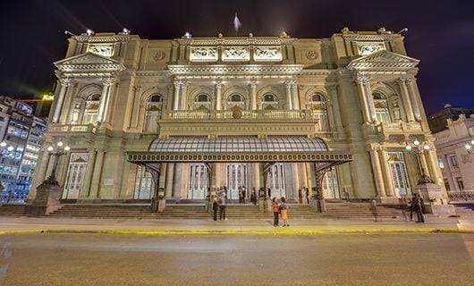 A grand, historic theater building illuminated at night, its ornate columns and large windows glowing like a total solar eclipse tour. A few people stand near the entrance on the wide steps leading up to the doors.