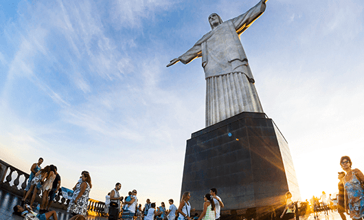 Tourists gather at the base of the Christ the Redeemer statue in Rio de Janeiro, Brazil, under a blue sky with the sun setting in the background, making it a memorable stop on their Solar Eclipse Tour through Brazil and Argentina.