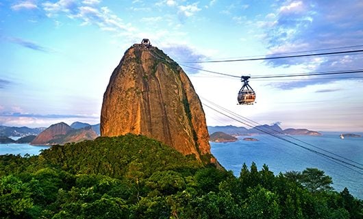A large, rocky mountain rises near the ocean in Brazil, with a cable car traveling up to its peak. Lush green trees cover the lower slopes as clouds drift above—a stunning sight for a Total Solar Eclipse Tour with views to Argentina in the distance.