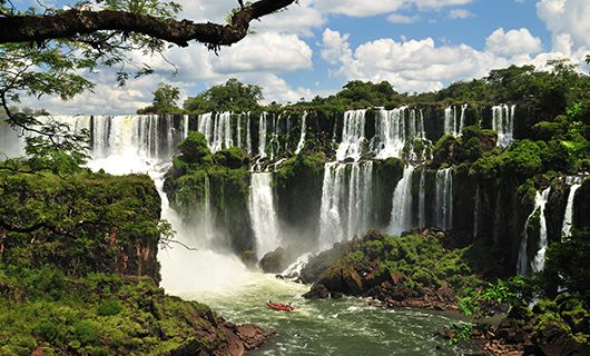 A wide view of Iguazu Falls on the Brazil-Argentina border with multiple cascading waterfalls surrounded by lush green vegetation, mist rising, and a small red boat in the river below under a partly cloudy sky.