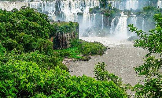 Lush green foliage frames a wide, powerful series of waterfalls cascading down rocky cliffs into a river below, with mist rising from the flowing water—an iconic sight straddling the border between Argentina and Brazil.