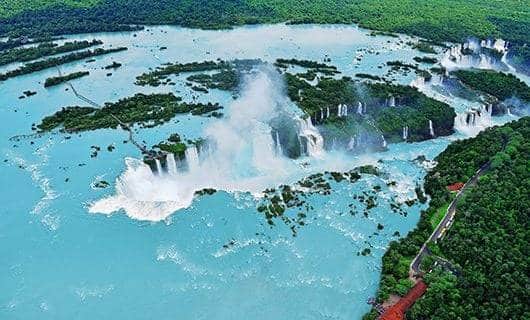 Aerial view of Iguazu Falls with multiple waterfalls cascading into turquoise water, surrounded by lush green rainforest and mist—an unforgettable stop on your Argentina solar eclipse tour.