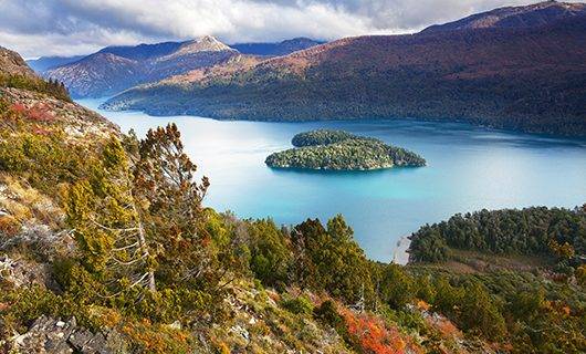 A scenic view of a turquoise lake surrounded by forested hills and mountains in Argentina, perfect for experiencing an Argentina solar eclipse, with a tree-covered island in the center and partly cloudy skies above.