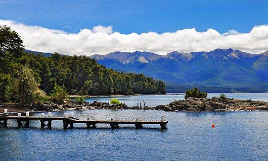 A wooden dock extends over a calm lake with forested shores and rocky outcrops—an ideal spot to experience a solar eclipse tour as tree-covered mountains rise in the background under a partly cloudy sky.