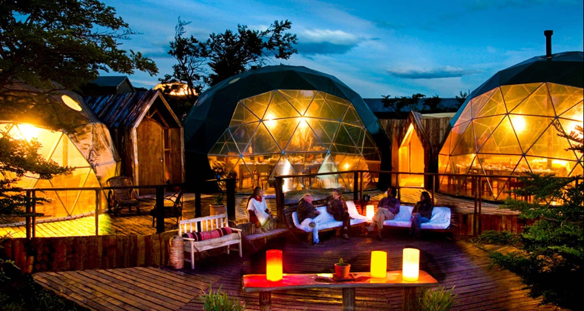 People relax on outdoor couches beside glowing candles, in front of illuminated geodesic domes and wooden cabins, under a twilight sky at this Patagonia Lodge. The setting combines modern comforts with nature, creating a cozy, inviting atmosphere.