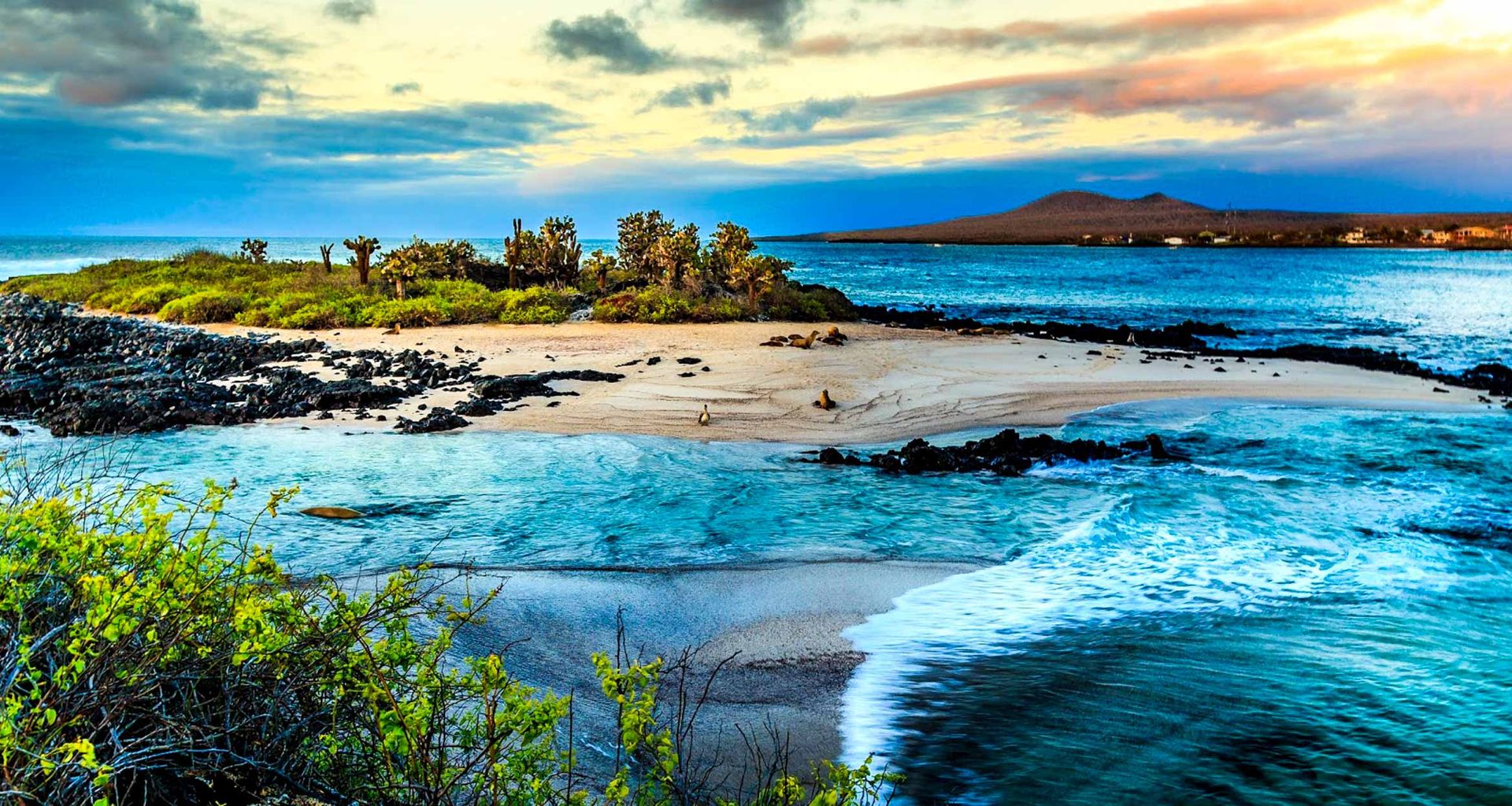 A scenic view of a small, sandy island surrounded by blue ocean waters, rocky shores, and lush vegetation under a partly cloudy sunset sky—perfect for a Galapagos cruise. In the distance, a larger landmass is visible.