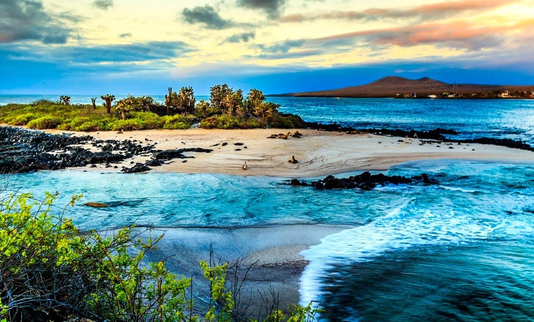 A small sandy island with rocky areas is surrounded by turquoise waves, dense green vegetation, and distant mountains under a colorful, cloudy sky at sunset—a perfect Galapagos tour setting.