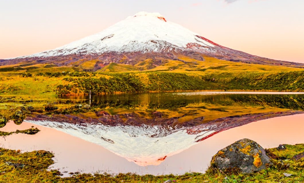 A snow-capped mountain reflected in a calm lake, surrounded by grassy hills and rocks under a pinkish sky at sunrise or sunset—an unforgettable view on your Ecuador Tour near Quito.