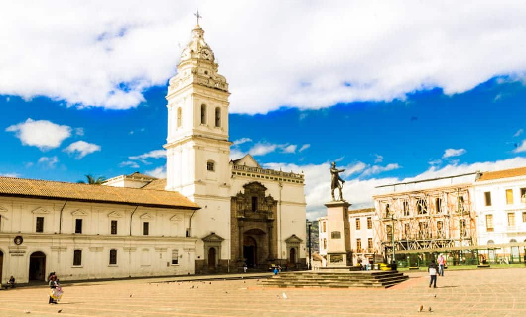 A sunny plaza in Quito with a white colonial church, bell tower, statue on a pedestal, and historic buildings under a blue sky. A few people and pigeons add life to the square—an iconic stop on any Quito Tour before heading to the Ecuador Amazon.
