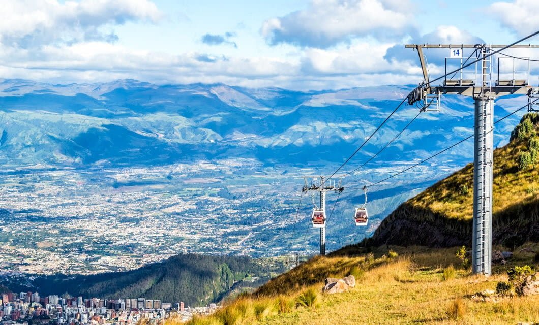 A cable car ascends a grassy mountain slope, overlooking a city nestled in a wide valley—evoking the dramatic green mountains and cloudy blue skies of Machu Picchu in the background.