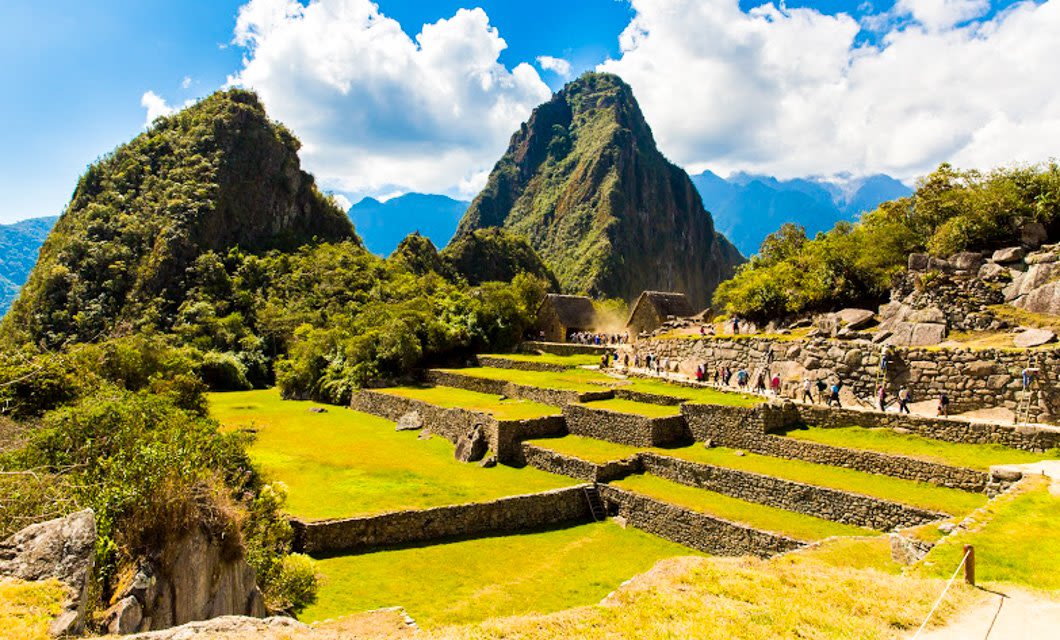 Wide view of Machu Picchu in Peru, showing ancient stone terraces, green grass, ruins, and lush mountains under a blue sky with scattered clouds—an iconic site like Easter Island, rich with history and mystery.