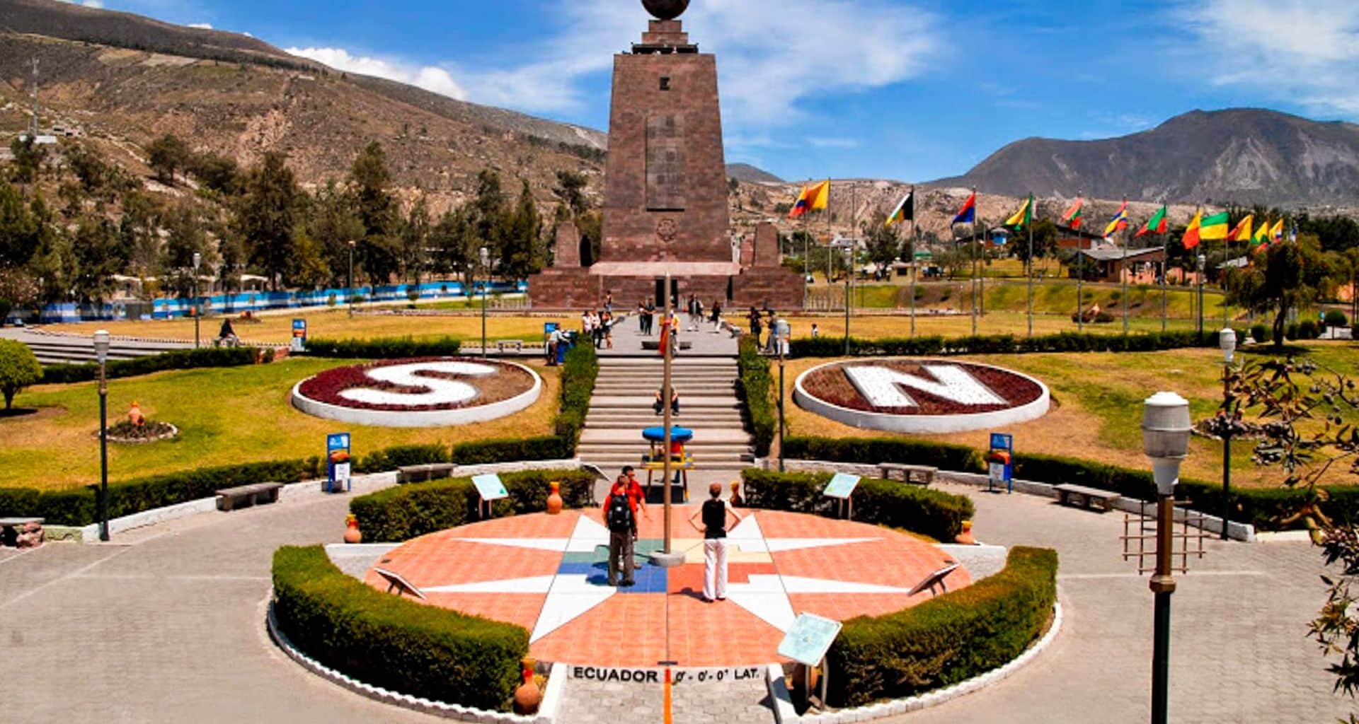 A monument marks the equator line in Ecuador, with a large globe on top. During a Quito tour, visitors stand on a star-shaped platform labeled with latitude, surrounded by gardens and flagpoles displaying country flags, with mountains in the background.