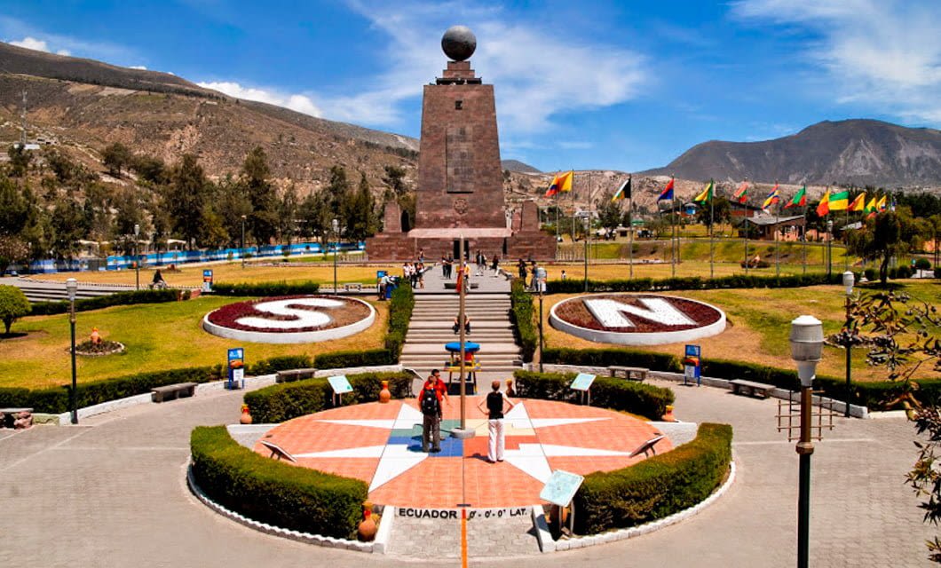 Tourists on an Ecuador Tour stand at the equator line in front of the Mitad del Mundo monument near Quito, with a globe atop the structure, gardens marked S and N, and mountains and flags in the background.