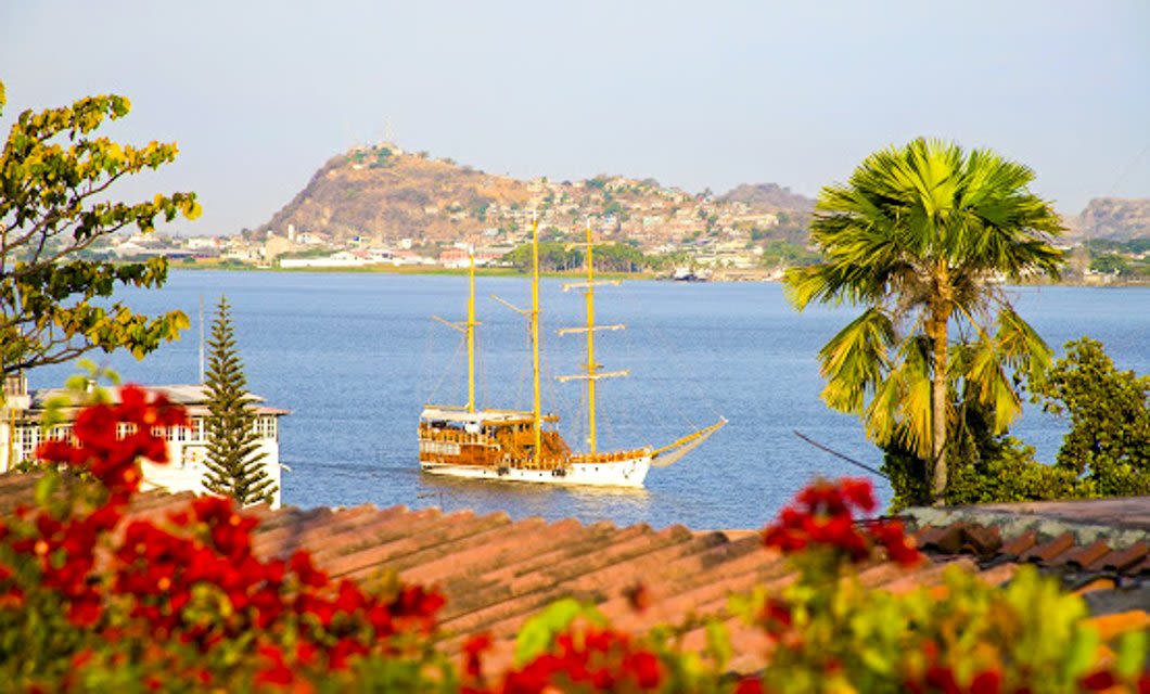 A wooden sailboat with tall masts floats on a calm blue lake, surrounded by red flowers and palm trees, as if waiting for travelers from a Tren Crucero train tour, with a hilly town visible in the background under a clear sky.