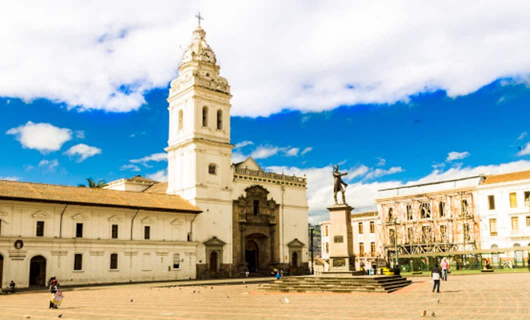 A large colonial plaza, reminiscent of Frederic Edwin Church’s Heart of the Andes painting, with a white church, bell tower, statue on a pedestal, and historic buildings under a bright blue sky. A few people and pigeons enliven the open square.