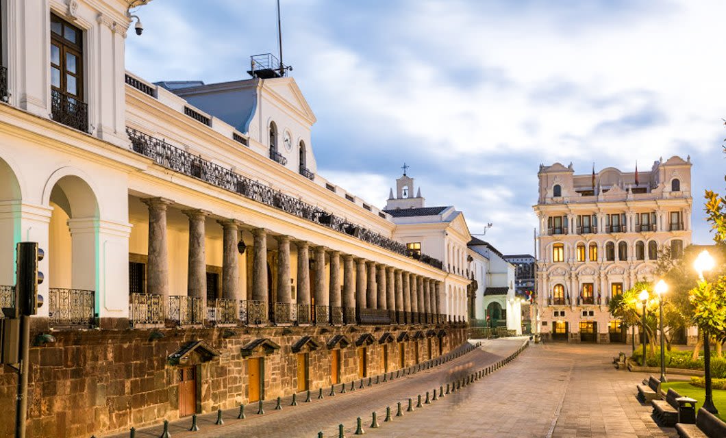 A wide, elegant colonial building with stone columns and arches stands beside a large plaza at dusk—a perfect scene to experience on a Quito tour, with another ornate white building and vintage street lamps glowing in the background.