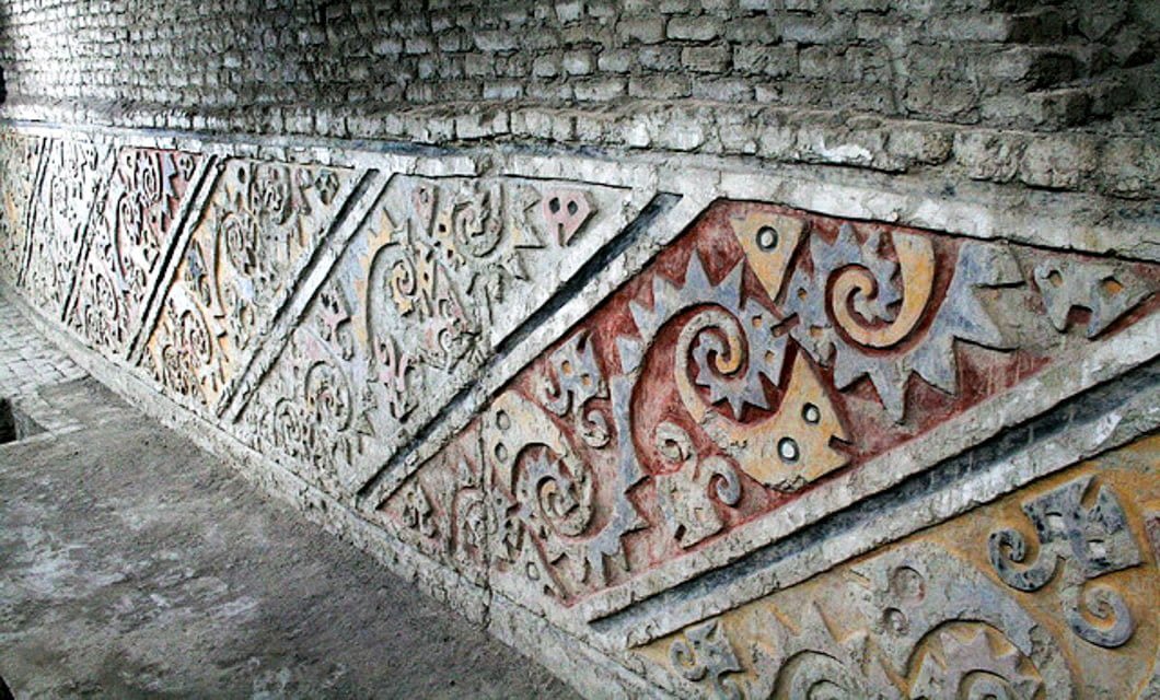Ancient adobe wall at Chan Chan featuring colorful geometric and swirling patterns in red, yellow, and white, located in an archaeological site near Trujillo with exposed brick above the decorated section.